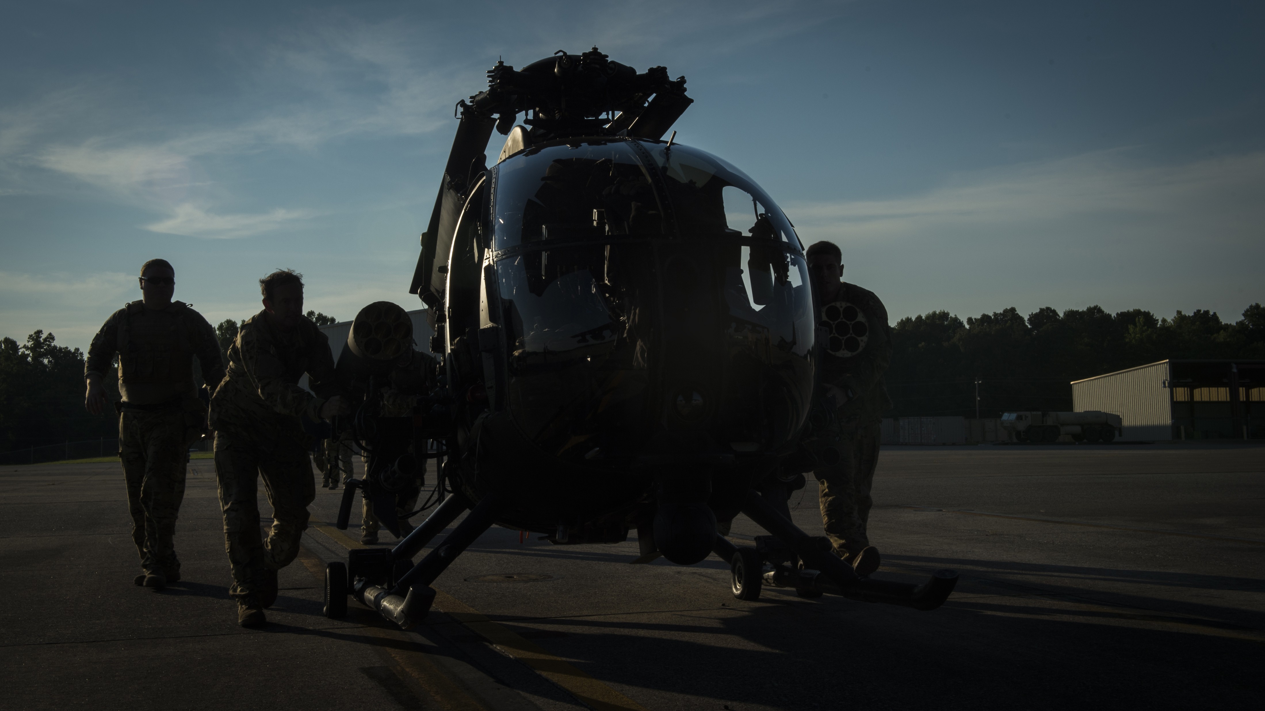 Dark AH-6 Little Bird on a ramp at low light with personnel gathered around the helicopter.