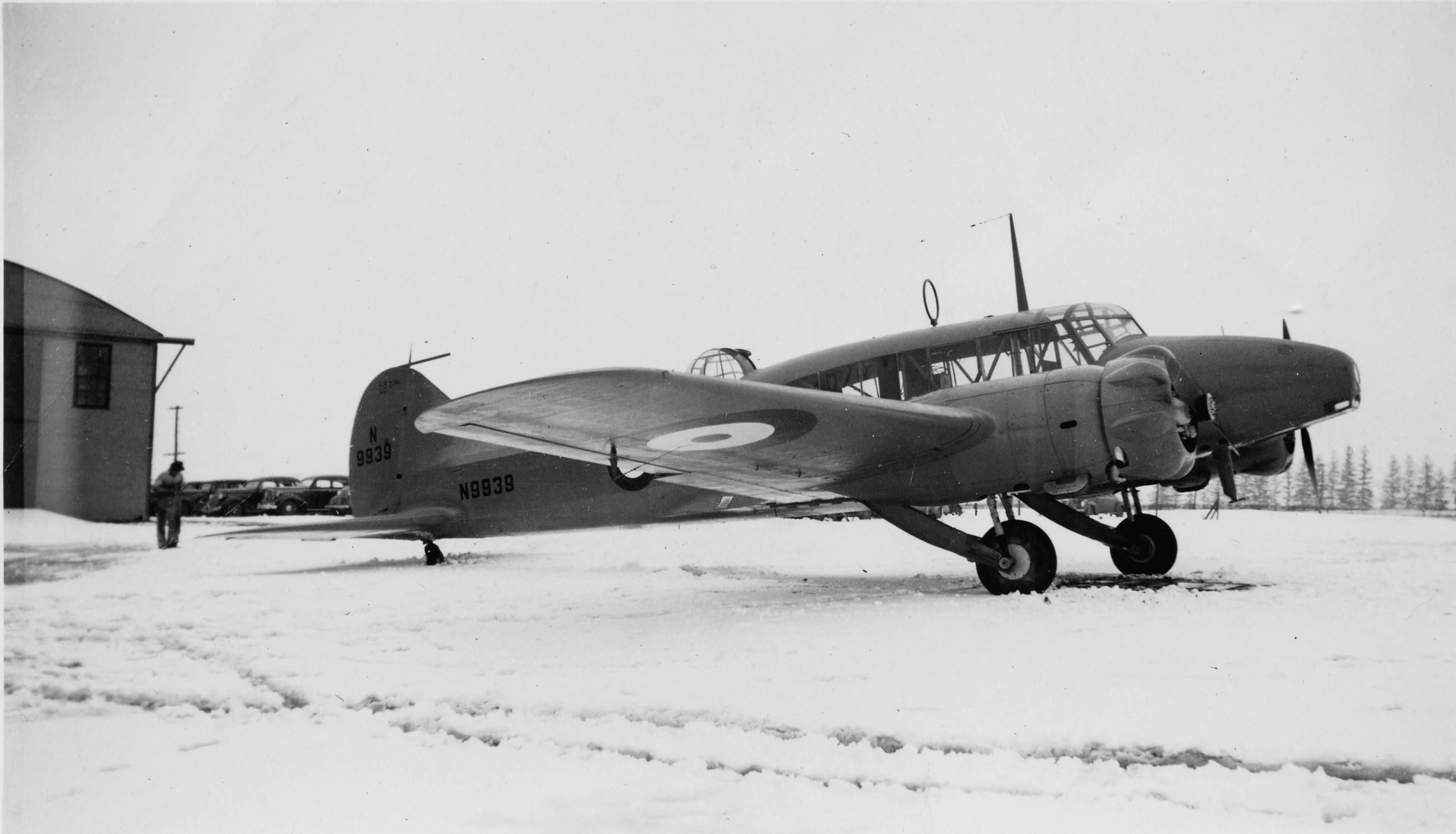 Black-and-white Avro Anson N9939 parked on snow beside a building.