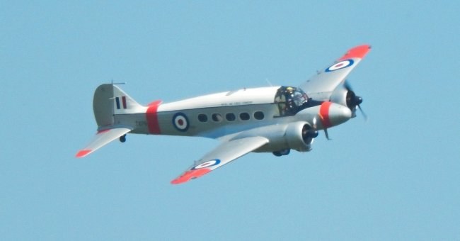 Repeat view of a silver Avro Anson flying with RAF markings and red trim.