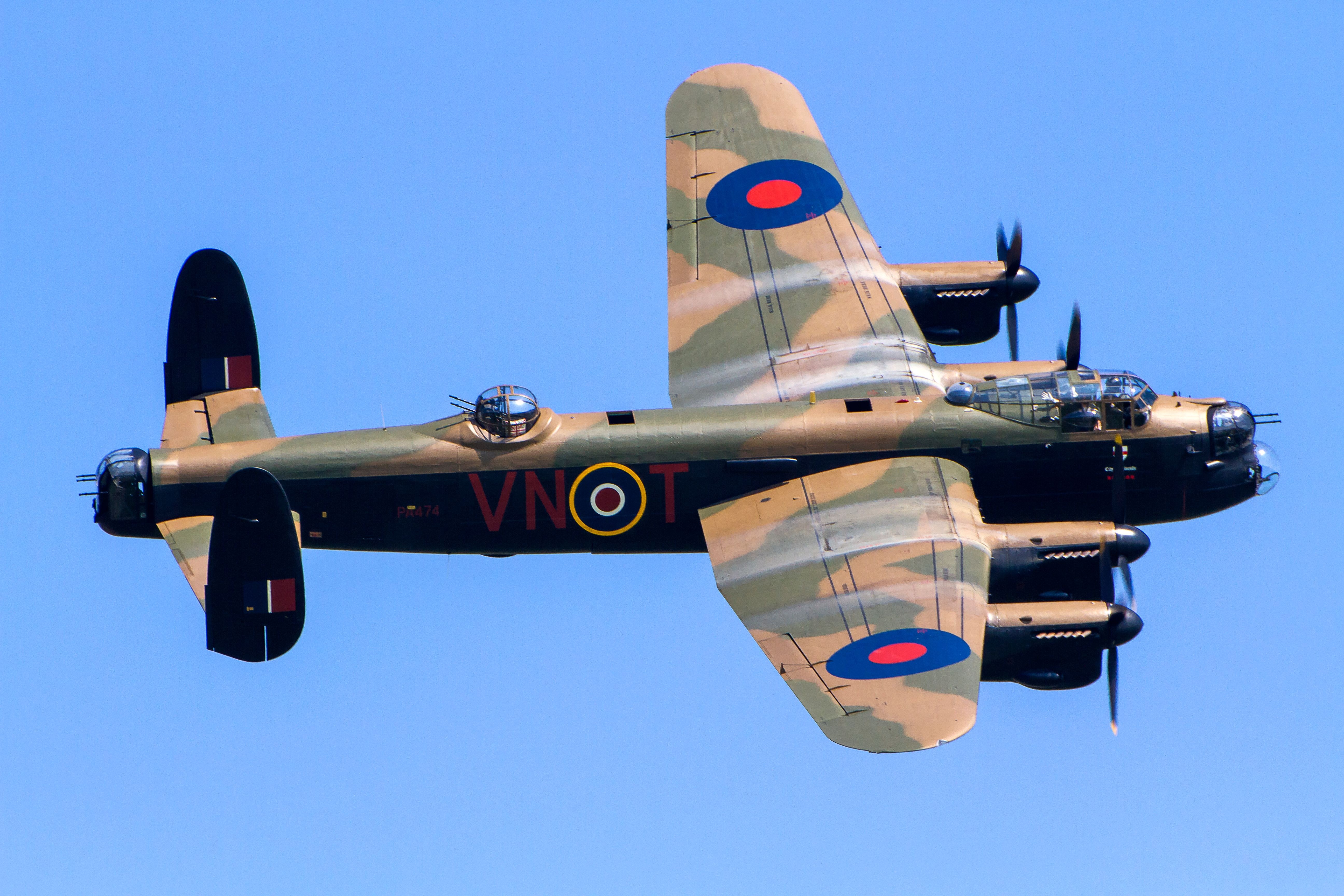 Color in-flight view of a camouflaged Lancaster showing upper wings, turrets, and RAF markings.