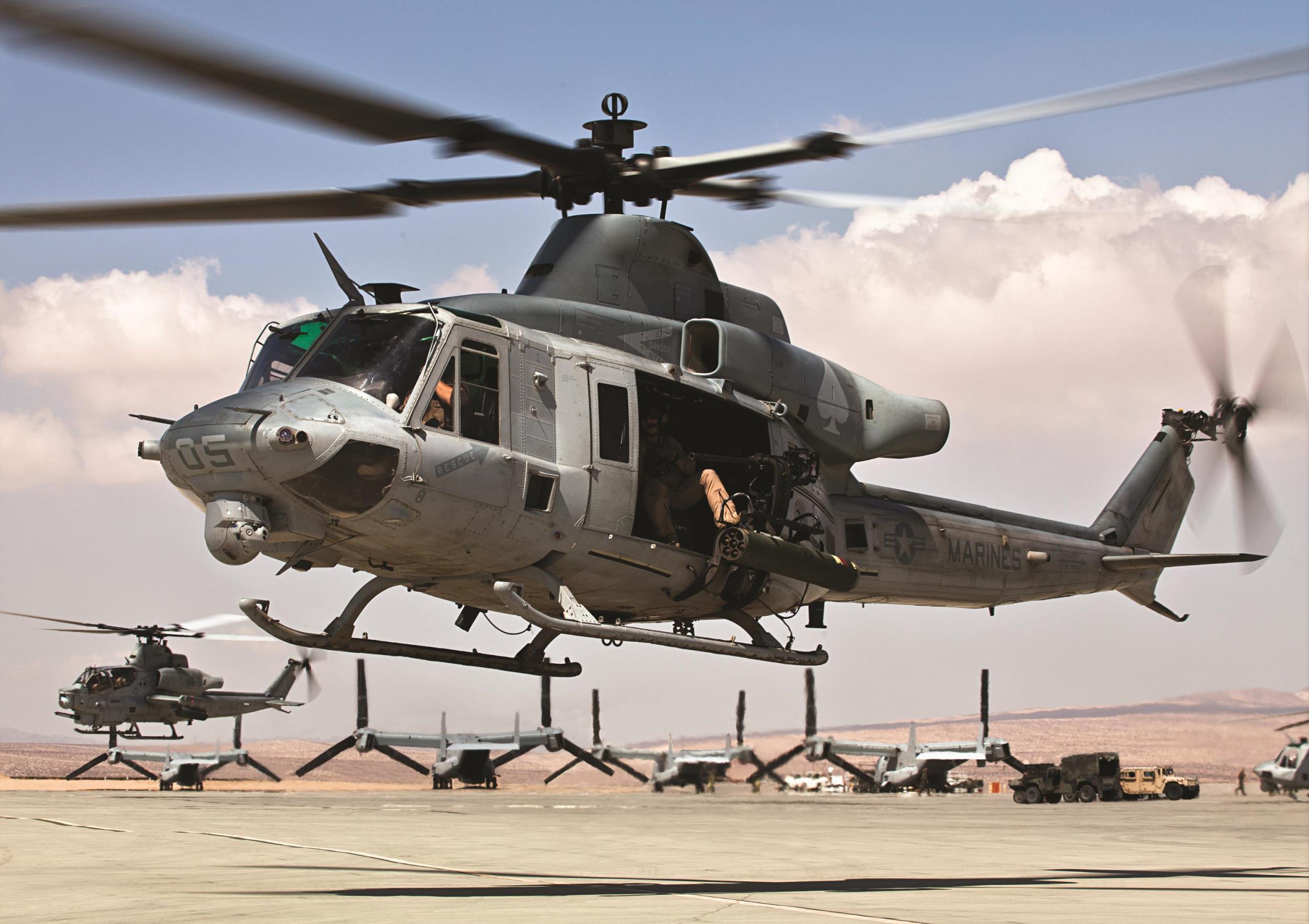 Marine Bell UH-1Y hovering near a flightline with other rotorcraft parked behind it.