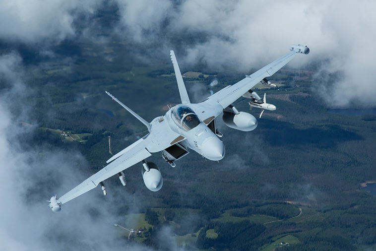 Gray Boeing EA-18G Growler flying above clouds and forested terrain.