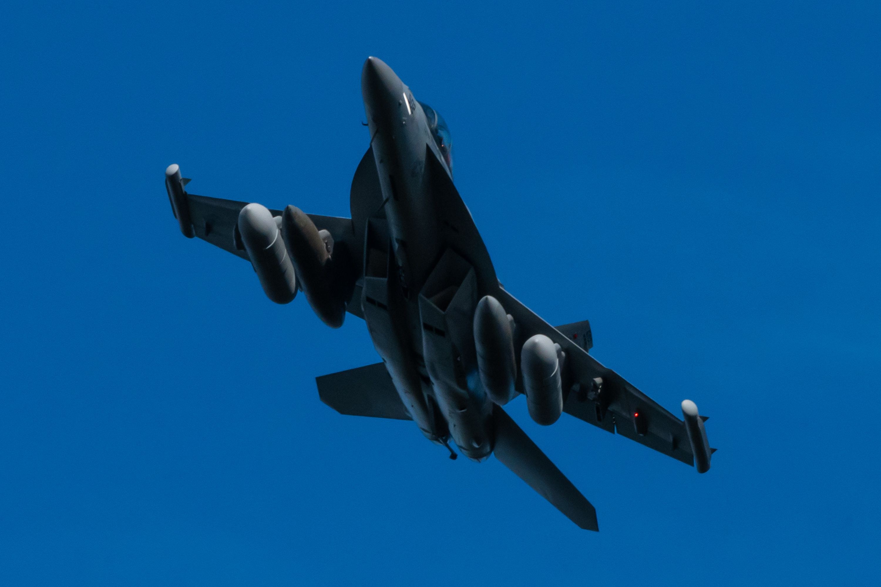 Dark underside view of an EA-18G Growler climbing against a blue sky.