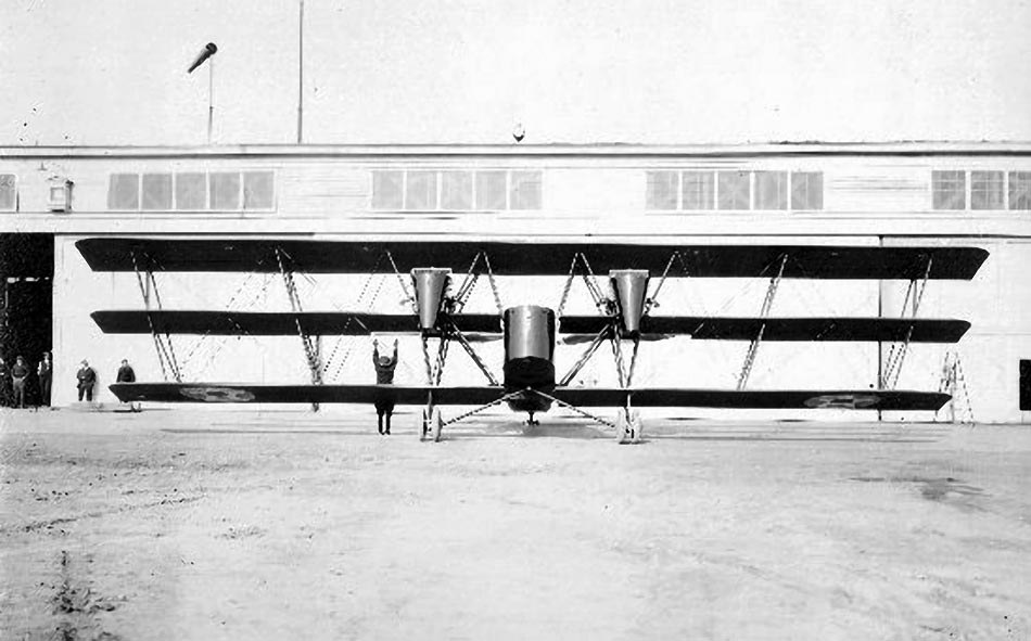 Boeing GA-1 triplane facing a hangar with a person under the wing.