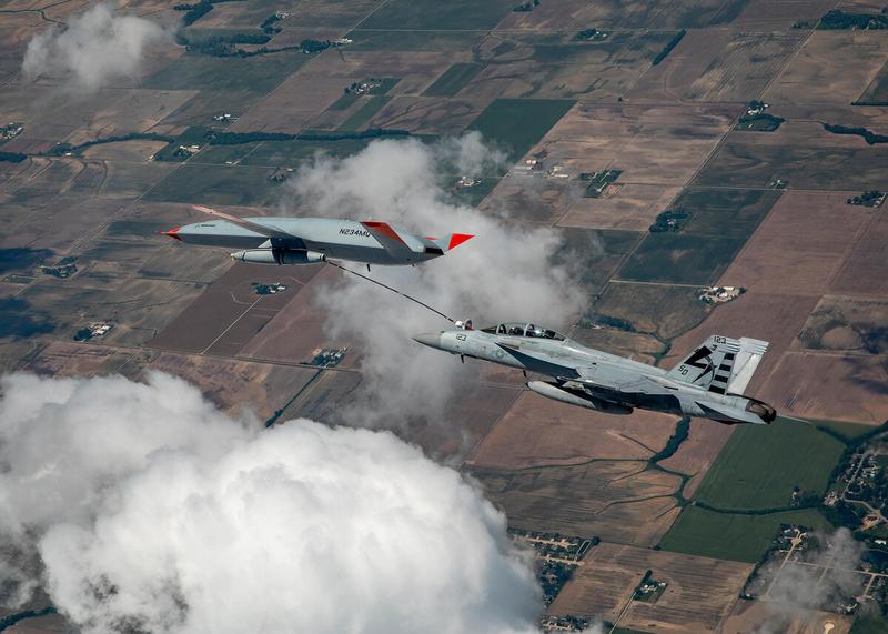 Boeing MQ-25 Stingray refueling a Navy F/A-18 over fields and clouds.
