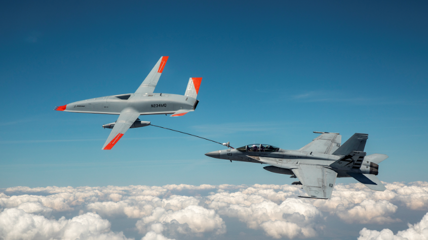 Boeing MQ-25 Stingray and Navy F/A-18 connected by a refueling hose above clouds.