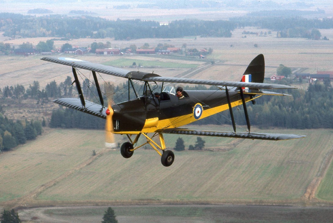 Olive-and-yellow de Havilland Tiger Moth flying above fields.
