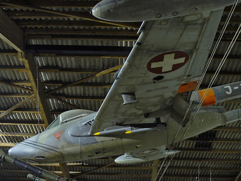 Grey de Havilland Venom suspended in a hangar with a Swiss cross under the wing.