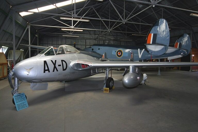 Silver de Havilland Venom marked AX-D displayed indoors with stores under the wing.