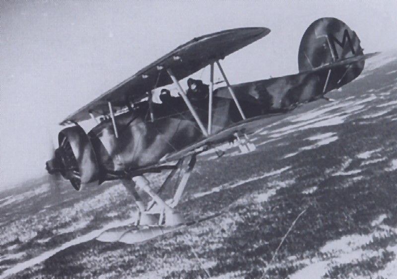 Black-and-white image of a dark biplane flying above snow-covered ground on skis.