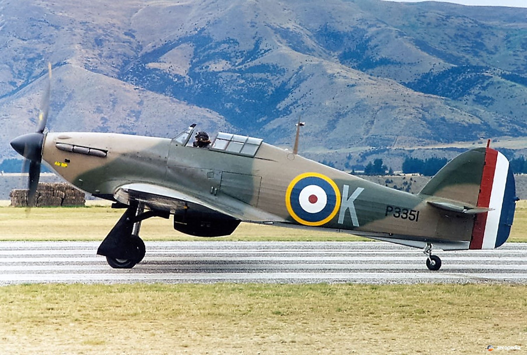 Camouflaged Hawker Hurricane P3351 taxiing on a runway with mountains behind it.