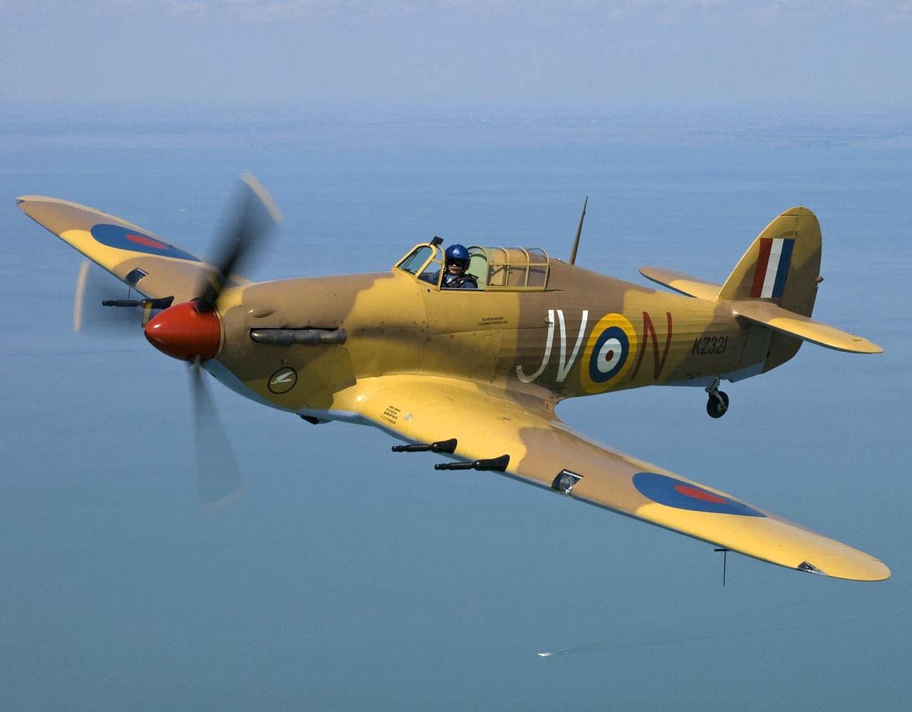Tan Hawker Hurricane KZ321 marked JV-N flying over open water.