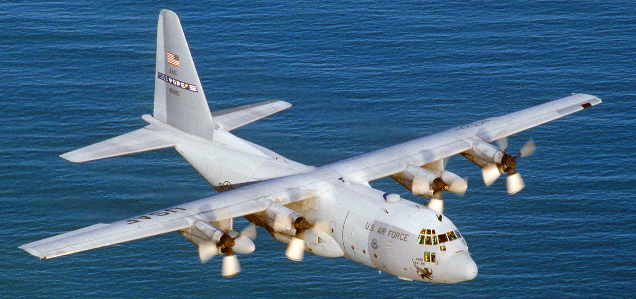 White U.S. Air Force Lockheed C-130 Hercules flying over water with Pope tail marking.
