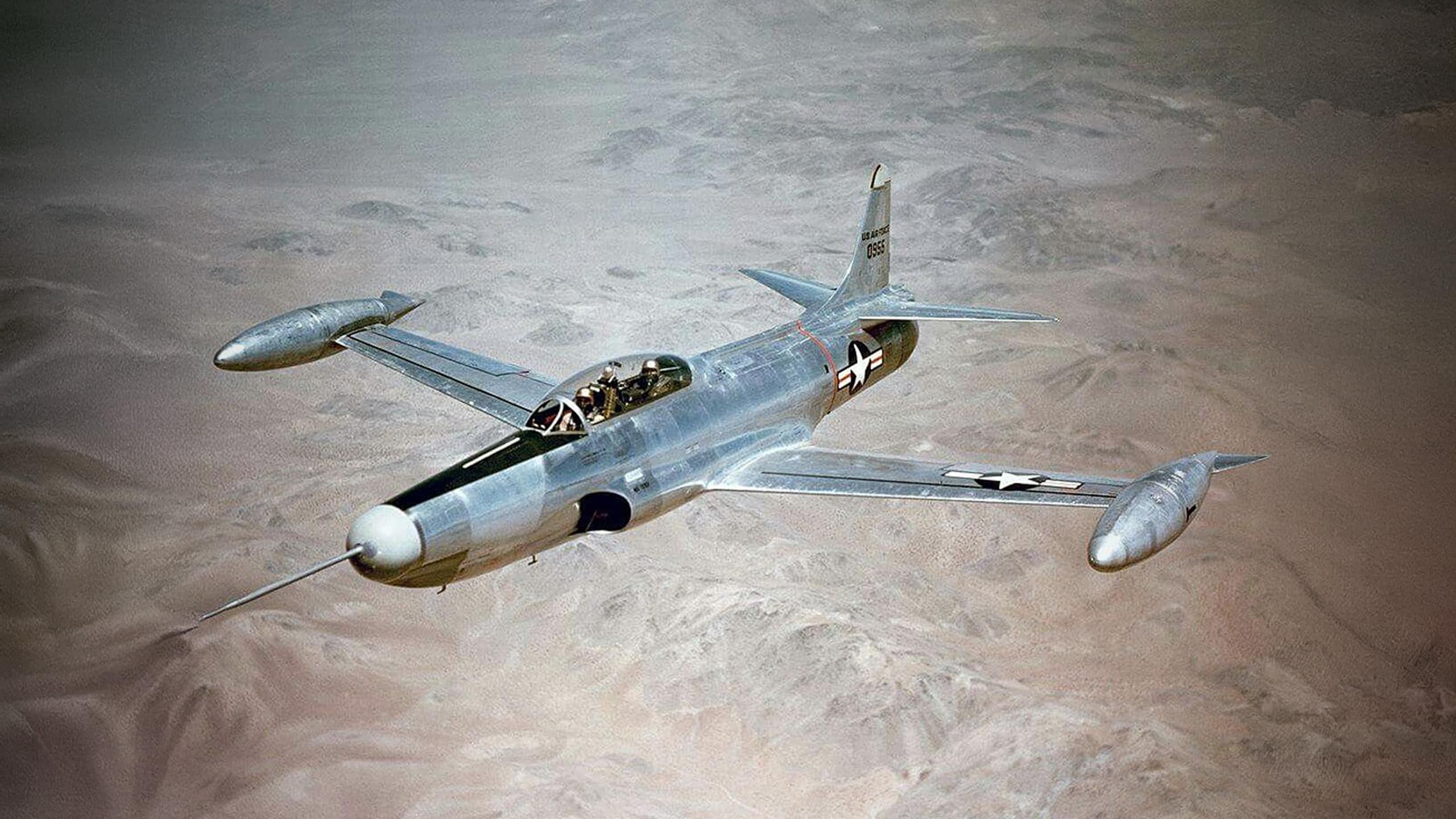 Silver F-94 Starfire flying over desert terrain with wingtip tanks.