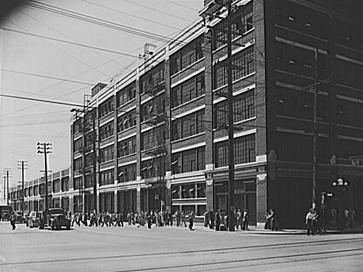 Black-and-white street view of the Lockheed aircraft plant in Burbank, showing the multi-story production building and vehicles outside the facility.