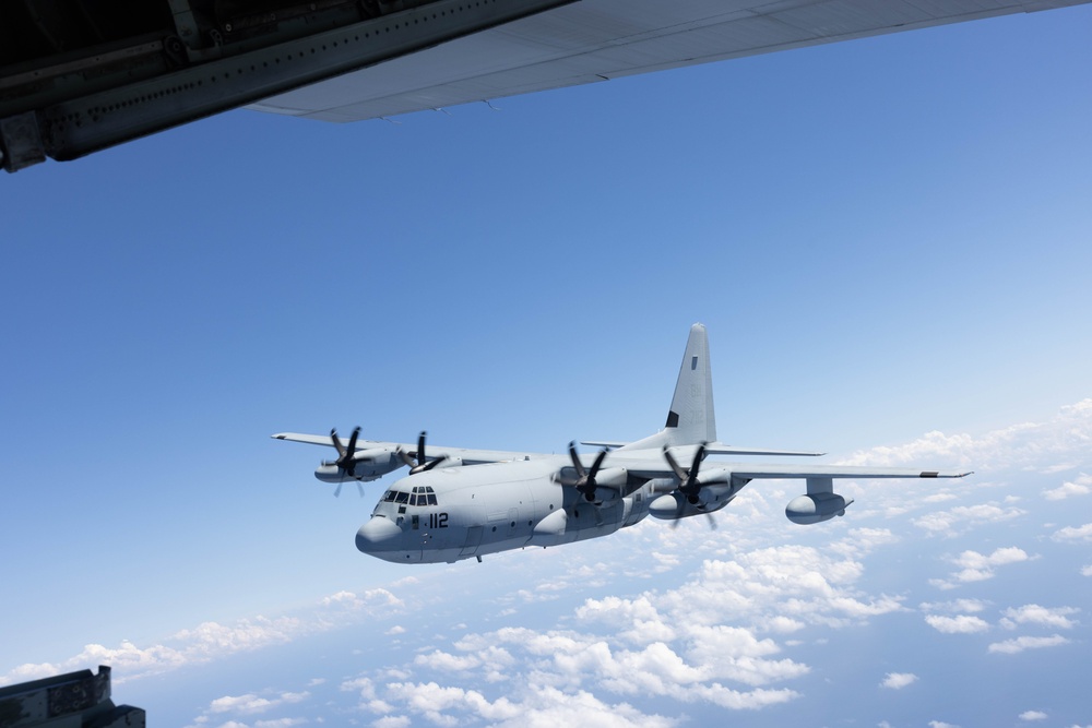 KC-130J 112 flying below another aircraft wing over clouds.