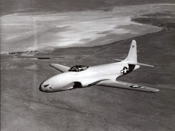 Black-and-white P-80 Shooting Star flying above an airfield.