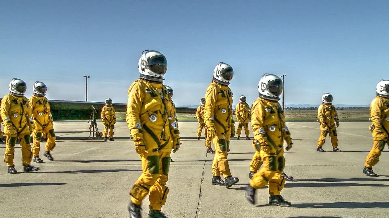 Personnel in yellow pressure suits walking on an airfield ramp.