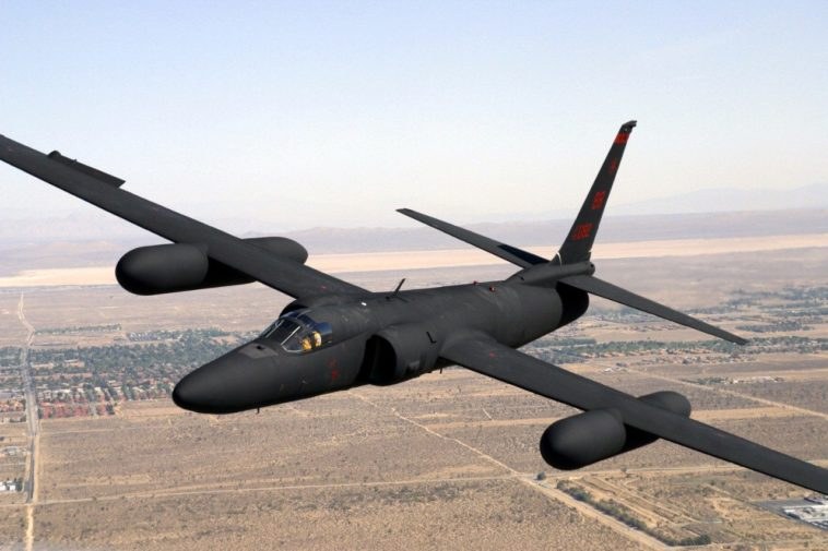 Black U-2 flying over desert terrain with long wings and underwing pods.