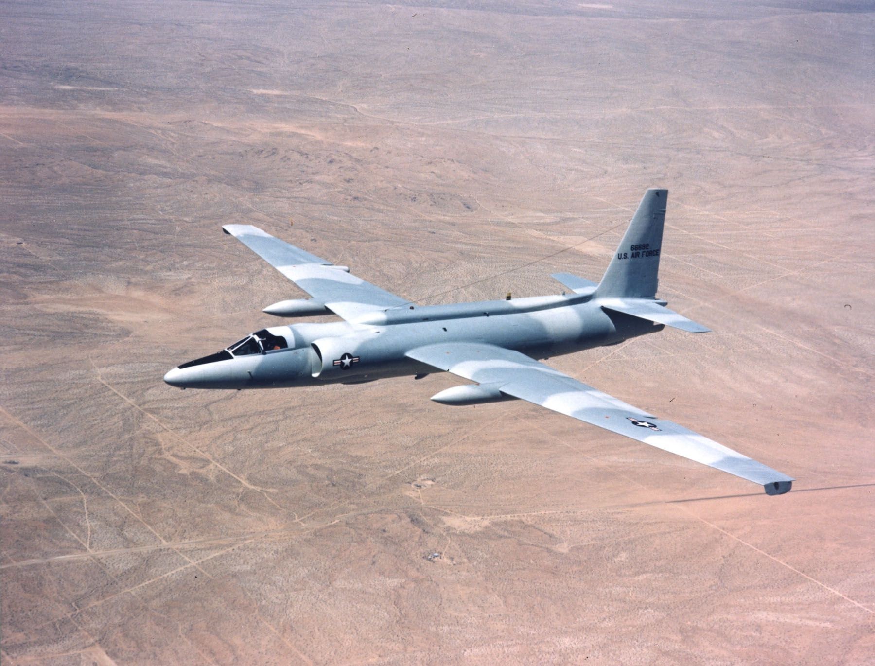 Silver U-2 flying over desert terrain with long wings and underwing pods.