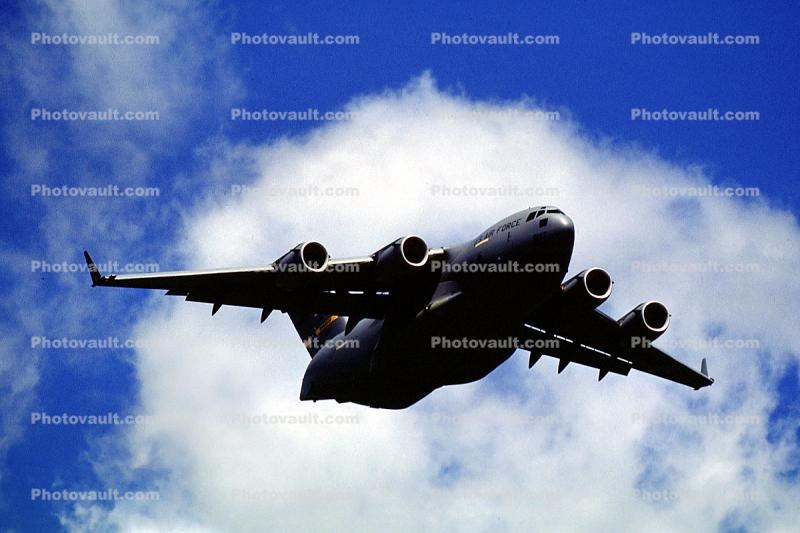C-17 Globemaster III flying over clouds with underside and engines visible.