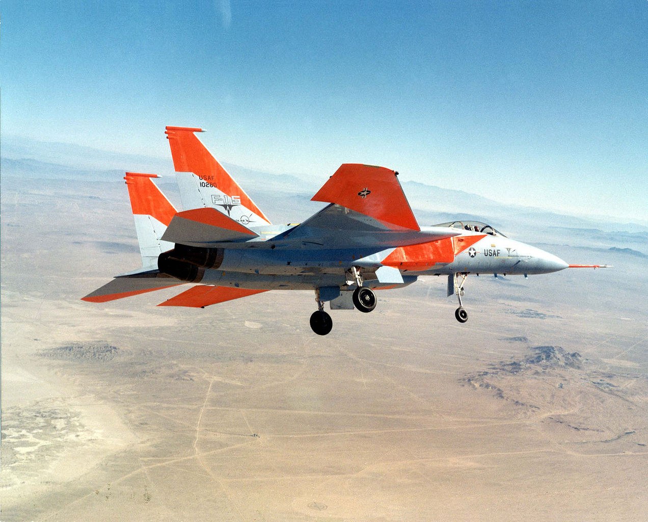 Orange-and-white F-15 Eagle flying over desert terrain with landing gear down.