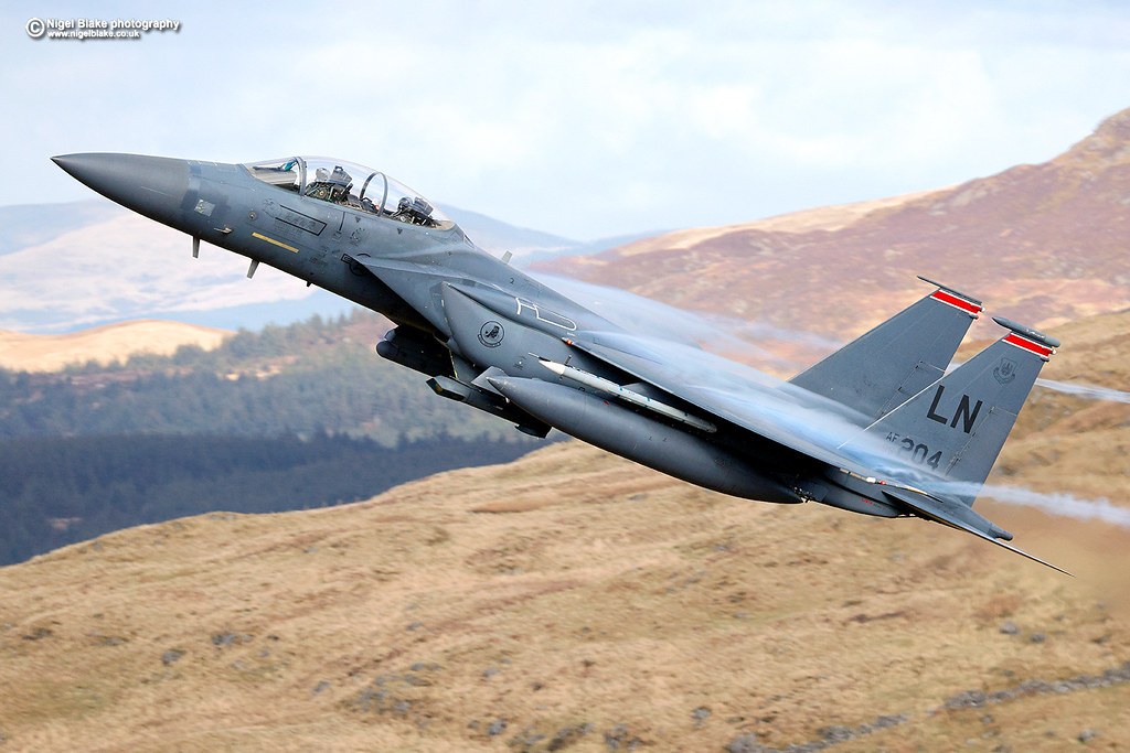 F-15E Strike Eagle marked LN 0204 banking above a hilly landscape.