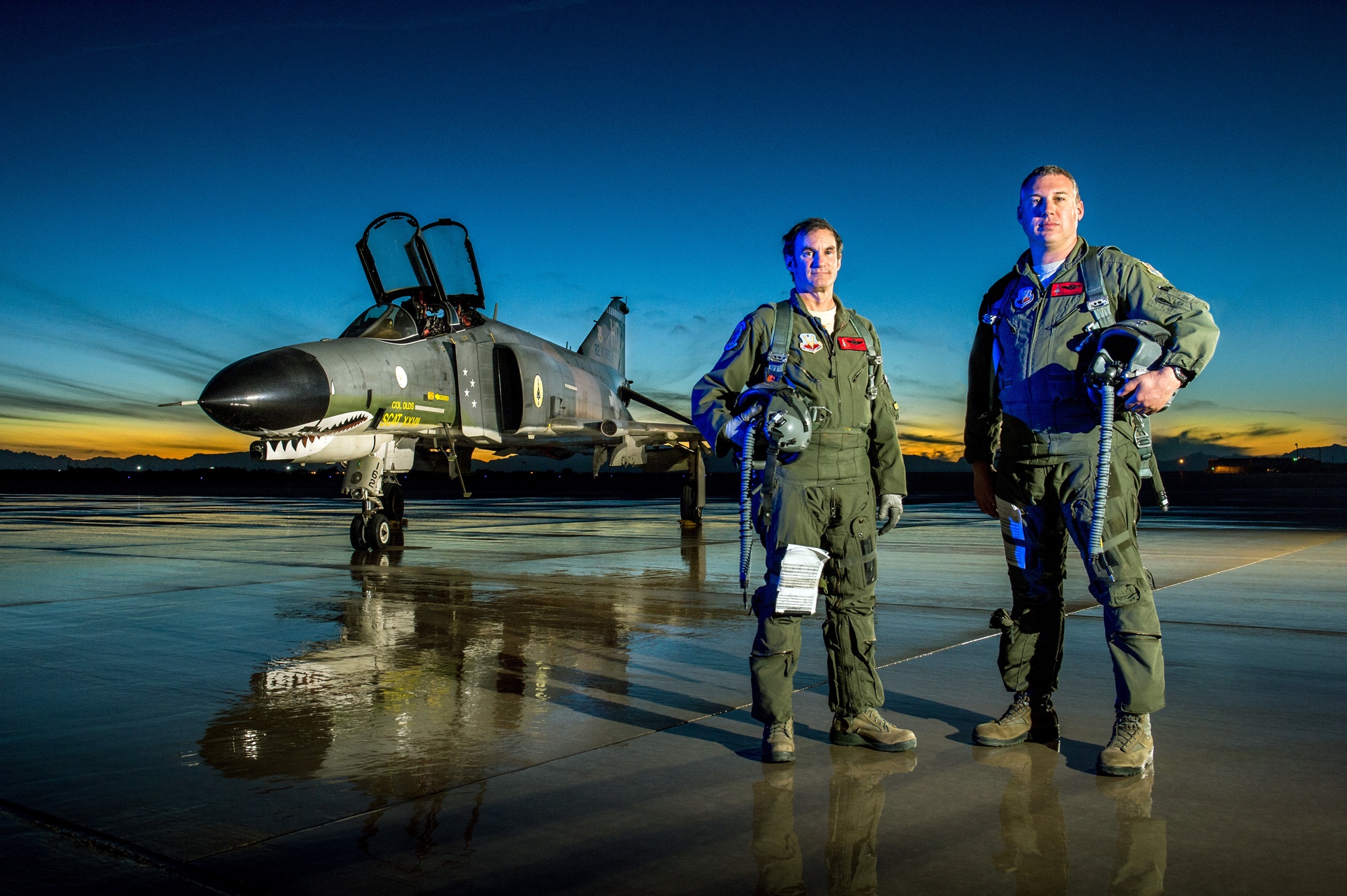 F-4 Phantom II with shark-mouth nose art parked on a wet ramp at dusk with two aircrew standing in front.