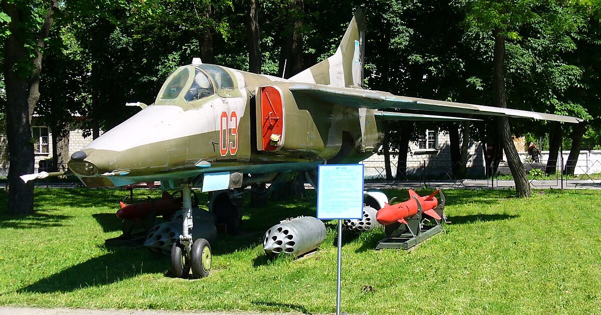 Camouflaged MiG-27 numbered 03 displayed outdoors on grass with stores nearby.