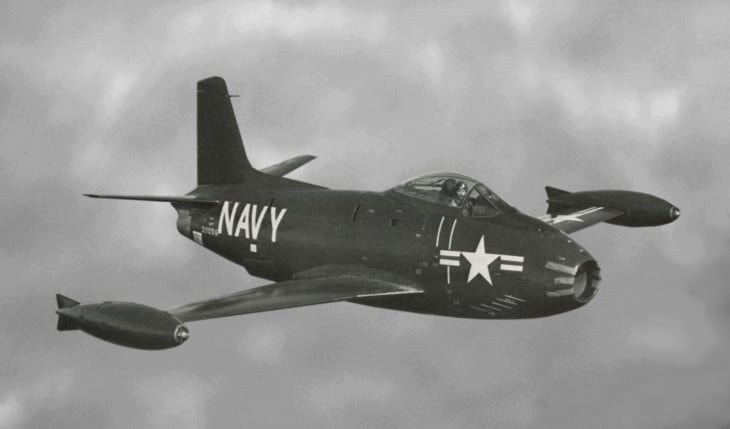 Black-and-white view of a Navy FJ-1 Fury flying with wingtip tanks and gear retracted.