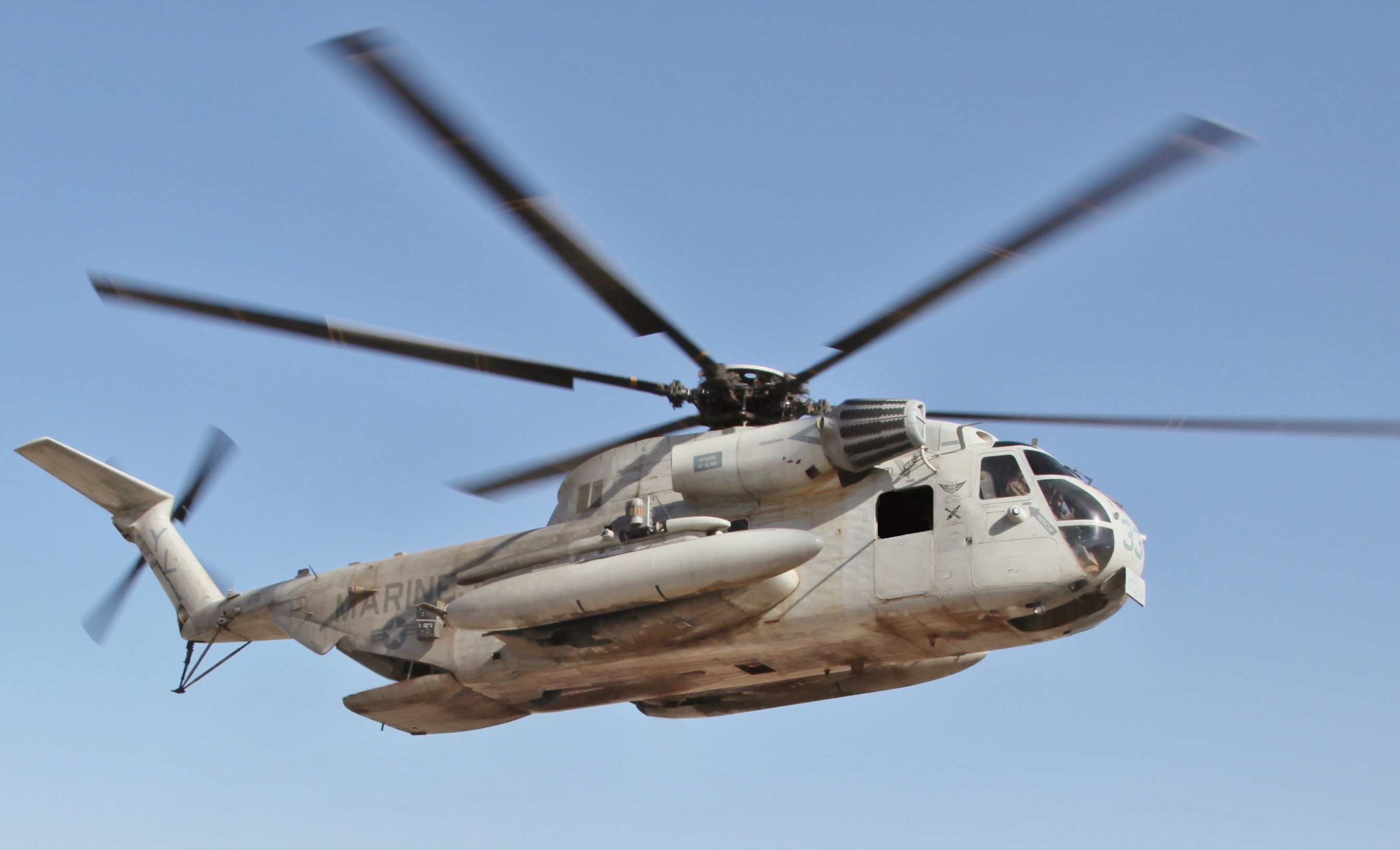 Light-colored Sikorsky CH-53 Sea Stallion flying against a clear blue sky.